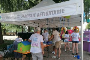 Persone che indossano camicie bianche stanno sotto un tendone con uno striscione che recita "COORDINAMENTO FAMIGLIE AFFIDATARIE", allestendo tavoli con palloncini, bottiglie d'acqua e materiale informativo durante un evento all'aperto.