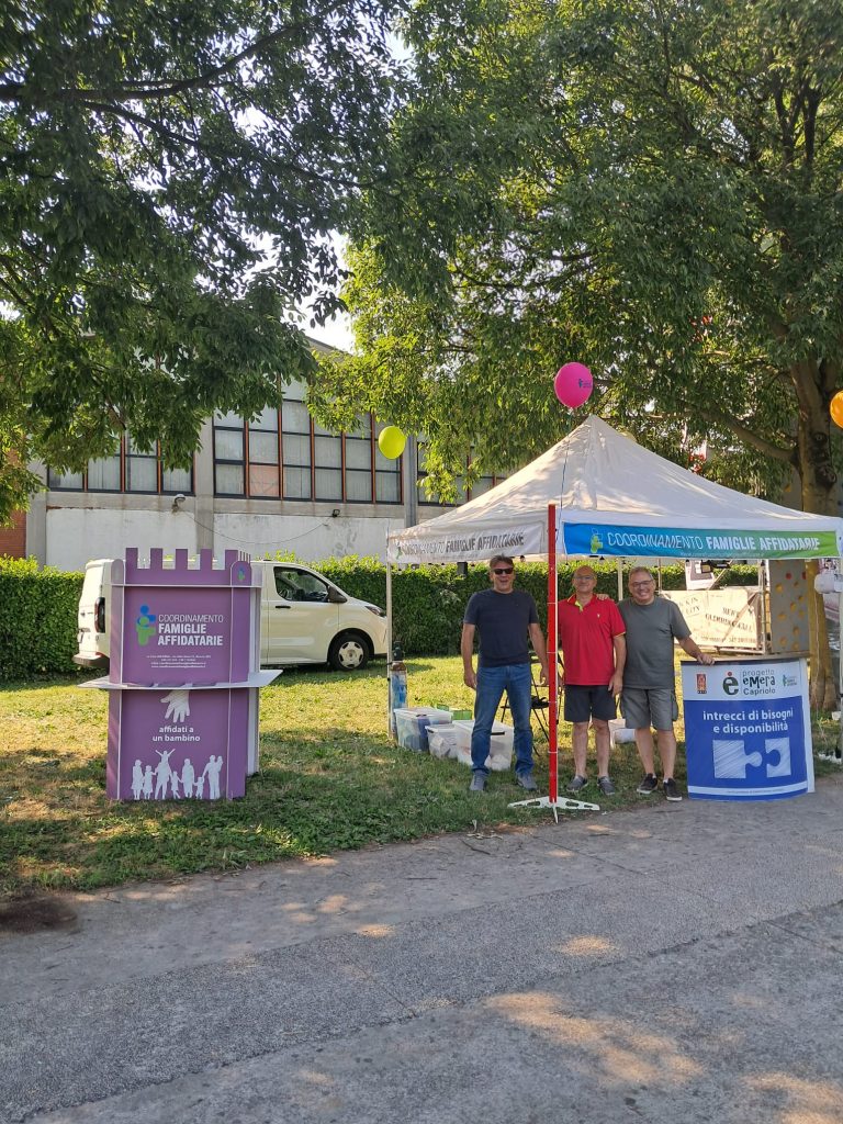 Tre persone sono in piedi e sorridono vicino a uno stand sotto una tenda bianca durante un evento all'aperto. Lo stand ha cartelli informativi sulle famiglie adottive. Sullo sfondo sono visibili alberi, erba, palloncini e un edificio.