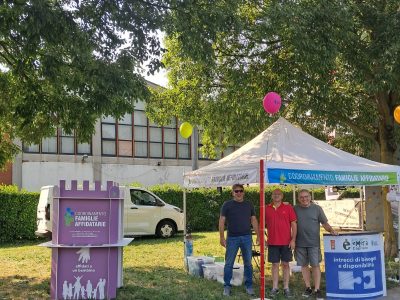 Tre persone sono in piedi e sorridono vicino a uno stand sotto una tenda bianca durante un evento all'aperto. Lo stand ha cartelli informativi sulle famiglie adottive. Sullo sfondo sono visibili alberi, erba, palloncini e un edificio.